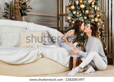 Lovely mom kissing her adorable daughter girl exchanging gifts on Merry Christmas and Happy New Year. Mother and little child having fun near fur tree. Christmas holiday concept. Copy ad text space