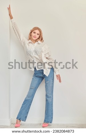 Full-length portrait of a cute blonde girl with delicate pink makeup posing in an elegant white blouse and jeans on a pink studio background. Hairstyles, Hollywood wave. Femininity. 