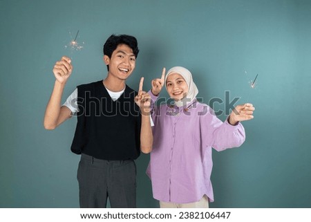 two young asian people are celebrating new year with pointing up side pose while holding firework in front of blue background