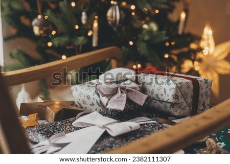 Stylish christmas gifts in festive wrapping paper with bows, vintage ornaments on chair on background of golden christmas star and tree lights. Happy holidays! Atmospheric christmas eve