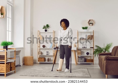 Young ethnic woman walking with crutches at home. African American girl with a broken leg walking with crutches in the living room. Accident, injury, fracture, treatment concept Royalty-Free Stock Photo #2382114453
