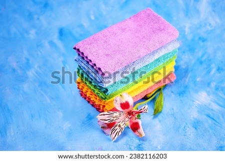 a spotted bright alstroemeria flower and seven rainbow-colored cleaning rags lie on top of each other for cleaning the room, on a blue background close-up Royalty-Free Stock Photo #2382116203