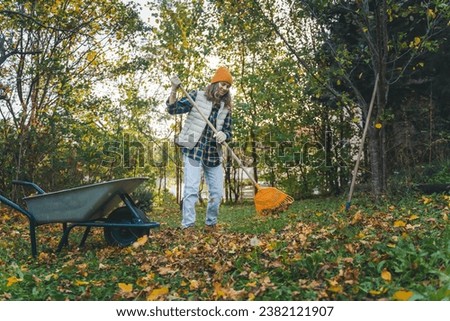 Young happy  woman collecting autumn leaves in the backyard of a country house using a rake and cart Royalty-Free Stock Photo #2382121907