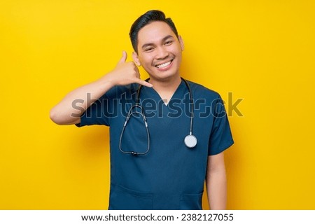 Smiling happy professional young Asian male doctor or nurse wearing a blue uniform and stethoscope showing gestures like saying call me back for consultation isolated on yellow background
