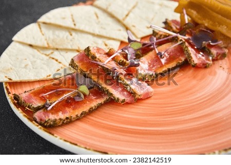 A close-up view of finely sliced tataki, beautifully seasoned and garnished with fresh microgreens. Accompanied by lightly grilled tortillas, it's presented on a vibrant orange plate. Royalty-Free Stock Photo #2382142519