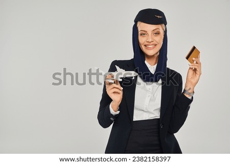 young arabian airlines stewardess in uniform holding airplane model and credit card on grey