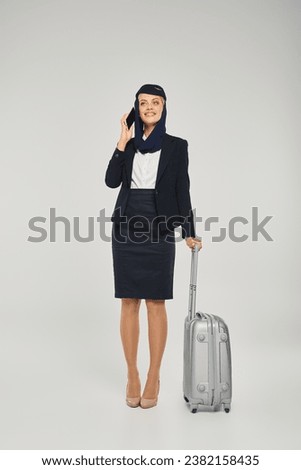 pleased arabian airlines air hostess with travel bag talking on mobile phone on grey backdrop