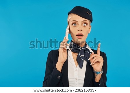 amazed air hostess in uniform talking on mobile phone and showing idea gesture on blue background