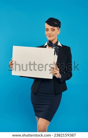 attractive stewardess in corporate uniform holding blank placard and looking at camera on blue