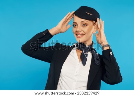 joyful woman in elegant uniform of air hostess adjusting elegant hat on blue background in studio