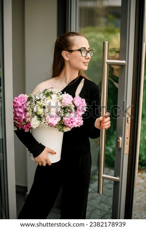 Attractive woman opens the door and holds in hand flower bouquet with hydrangea, peonies, carnations in white hat box. Flowers arragement created by florist