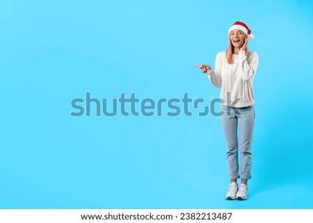 Delighted young woman in Santa hat and comfy white sweater exuberantly points aside at free space posing against striking blue background, full length