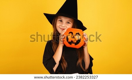 little girl with long hair smiles in a Halloween witch costume and holds a pumpkin-shaped candy bowl on a yellow background.
