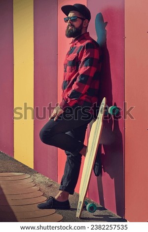 Full body of cool male skater in street style outfit standing with longboard against colorful striped wall on sunny day during free time