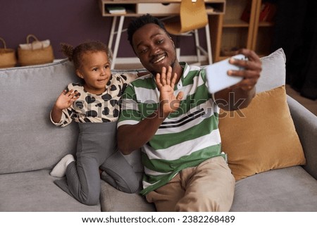 Portrait of happy Black father taking selfie photo with little daughter at home and waving Hello to camera