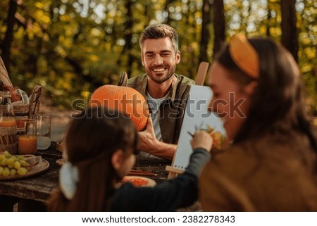 Caucasian mother and daughter are on a picnic in a forest during fall painting a picture, and the father is holding a pumpkin