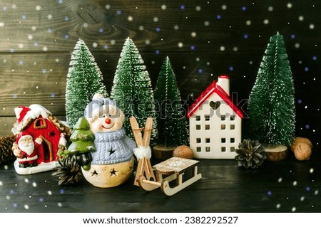 Christmas card, porcelain toy snowman with a Christmas tree in his hands and a New Year's forest house, green Christmas trees and a wooden sleigh.  Home decoration.  Background picture.