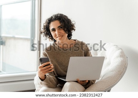 Happy young business woman using laptop, holding mobile phone, looking at camera in modern house. Portrait of successful lady sitting at home. Technology concept 
