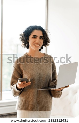 Smiling young business woman using laptop, holding mobile phone, looking at camera in modern house. Portrait of successful lady posing at home. Technology concept 