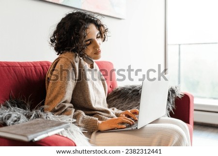 Busy middle eastern woman sitting at sofa and using smartphone while working with laptop at home. People and technology concept
