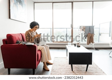 Curly middle eastern lady sitting at sofa and using smartphone while working with laptop at home. People and technology concept