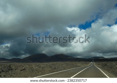 Long straight road in Lanzarote between volcanic rocks, dramatic sky with dark clouds. Volcanoes on the horizon.