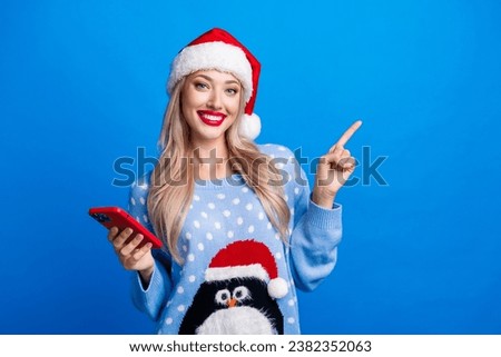 Photo portrait of lovely blonde young lady gadget point empty space dressed penguin sweater x-mas hat isolated on blue color background