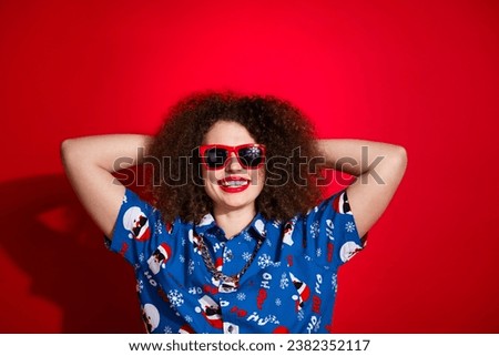Photo of pretty cheerful lady dressed ugly santa claus print xmas shirt arms behind head empty space isolated red color background