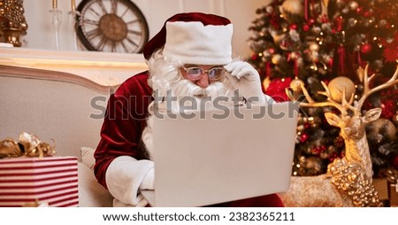 Santa Claus sitting at home and reading email on laptop with New Year wish list near Christmas tree