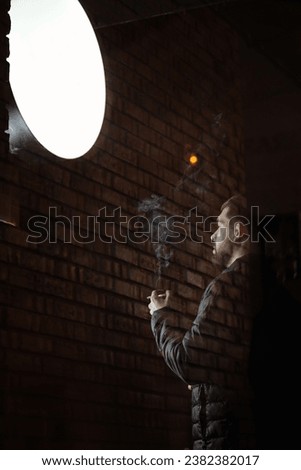 man with beard is enjoying smoking cigarette outdoors after hard work day on dusk by brick wall. view behind glass door. Cigarettes help with everything from boredom to anger management