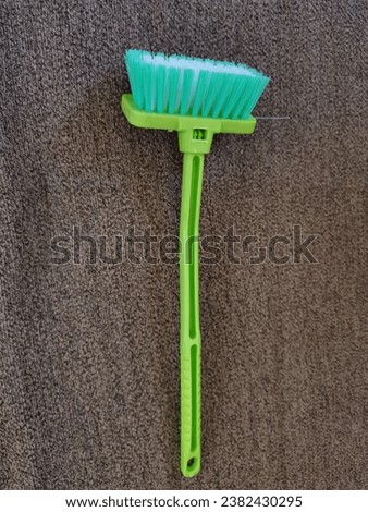 The new bathroom floor brush, green with white and blue bristles, is on the brown sofa, will soon be used to clean the bathroom. The photo was taken during the day.