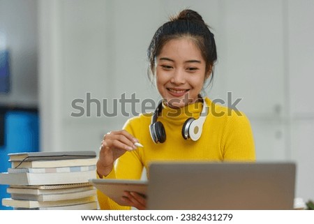 Happy Asian female student wearing wireless headphones studying online, learning, listening to lectures, browsing websites, searching for information on tablet and laptop in business study office.