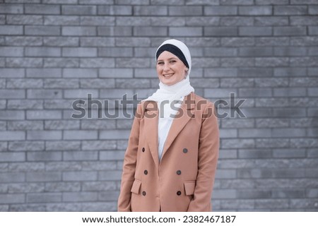 Portrait of a young blue-eyed woman in a hijab against a gray brick wall. 