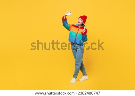 Full body side view young woman wears padded windbreaker jacket red hat casual clothes do selfie shot on mobile cell phone post photo on social network show v-sign isolated on plain yellow background