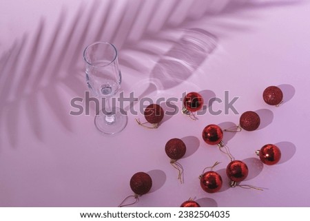 New Year party background with palm shadow, glasses, and red Christmas baubles on a pink pastel background. Christmas celebration and New Year idea. Flat lay.