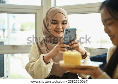 Young Muslim woman using mobile phone and taking photos of her friend holding cake.