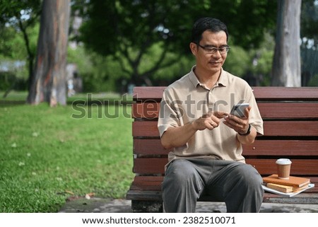 Image of carefree middle age man wearing glasses using mobile phone in the public park.
