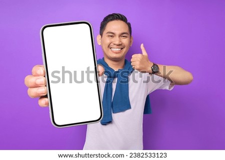 Cheerful young Asian man demonstrating smartphone with empty screen, showing thumb up gesture isolated on purple background. Advertising new mobile app, mockup concept Royalty-Free Stock Photo #2382533123