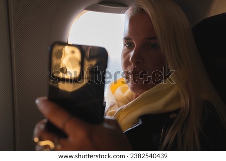 A blonde girl takes a selfie on her phone on an airplane while sitting at the window. High quality photo