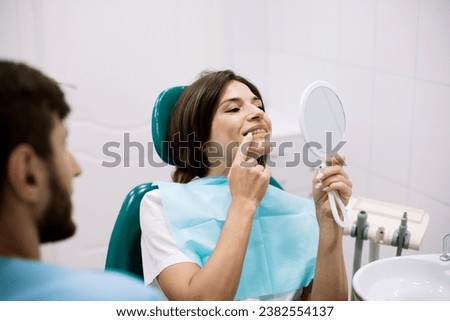 Young female patient holding mirror and looking at his beautiful smile sitting at the dental office Royalty-Free Stock Photo #2382554137