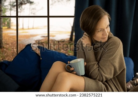 Pretty young woman reading book and drinking morning coffee at home sitting at the window behind which there is an autumn landscape