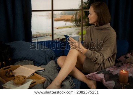 Pretty young woman reading book and drinking morning coffee at home sitting at the window behind which there is an autumn landscape Royalty-Free Stock Photo #2382575231