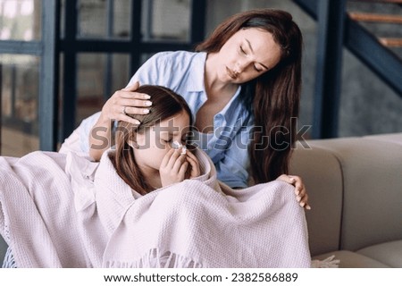 Sick child girl sitting on couch covered with blanket blowing nose, sneezing in tissues spending day at home with mother. Supportive mom hugging her ill daughter with flu symptoms Royalty-Free Stock Photo #2382586889