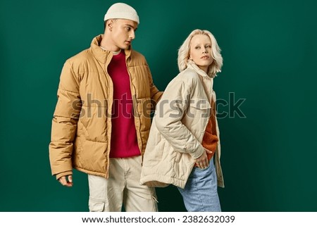 young couple in puffer jackets posing together on turquoise background, fashionable man and woman