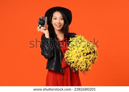 Young Asian woman with photo camera and chrysanthemum flowers on orange background