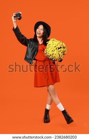 Young Asian woman with photo camera and chrysanthemum flowers on orange background