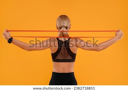 Woman exercising with elastic resistance band on orange background, back view