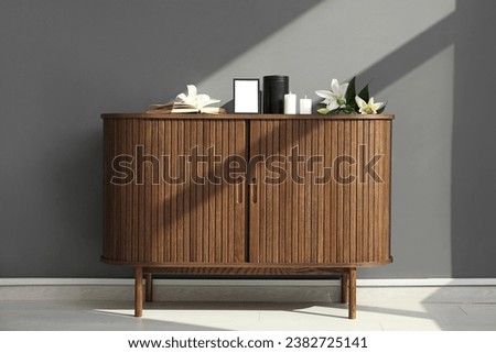 Blank funeral frame, burning candles, mortuary urn and lily flowers on wooden cabinet against grey wall