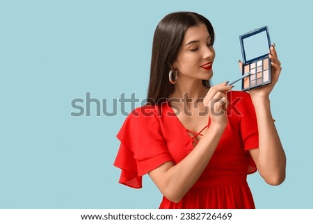 Beautiful young woman with brush and palette of eyeshadows on blue background