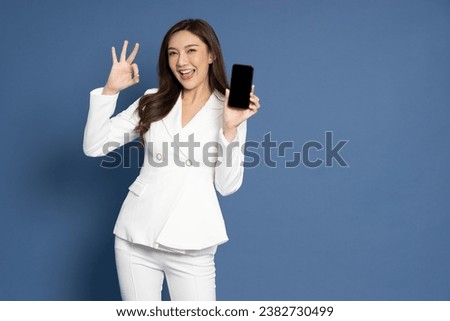 Portrait of Asian business woman in white suit showing or presenting mobile phone application and ok sign isolated over blue background, Asia Thai model